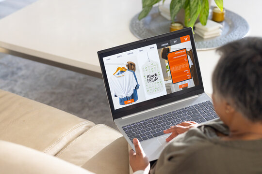 Mature African American woman sitting on beige leather sofa, browsing Black Friday sale on laptop