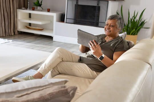 Senior African American woman reclining on sofa holding tablet smiling in living room, copy space
