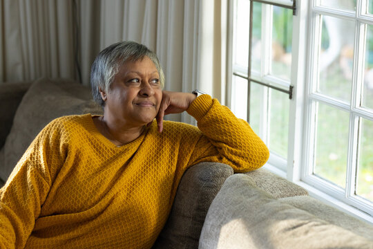 Senior woman sitting on gray sofa at home by window wearing mustard sweater watch gazing out