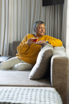 Senior African American woman sitting on sofa at home holding teacup with smartwatch looking right
