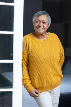 Senior African American woman standing smiling wearing mustard sweater by white glass door, lamp