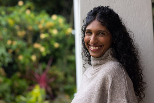 African American woman leaning on pillar at door, smiling, wearing sweater near flowers, copy space
