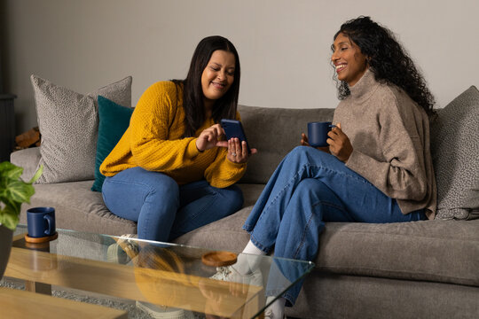 African American female friends sitting on gray sofa at home, tapping smartphone and holding mugs