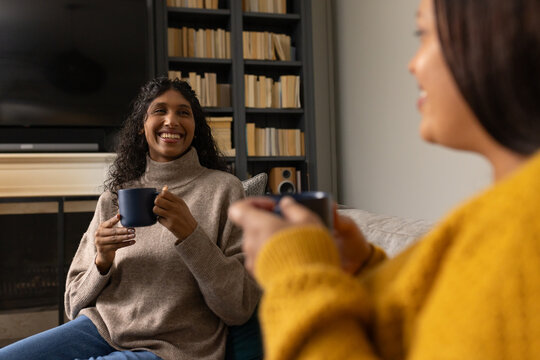 African American women in sweaters sitting on sofa in living room holding dark mugs and talking