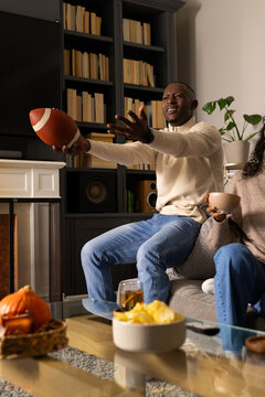 African American couple reacting and reaching on sofa in living room, man holding football
