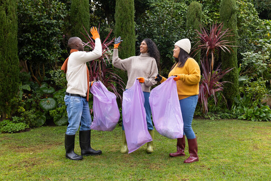 Diverse volunteers high-fiving while collecting lilac trash bags in garden with orange gloves