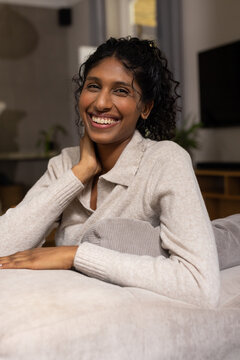 Woman wearing knit sweater sitting on light-colored sofa at home leaning on grey cushion smiling