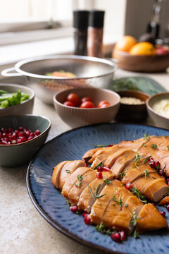 Glazed protein sitting on blue plate, getting thyme sprigs and pomegranate seeds, bowls near window