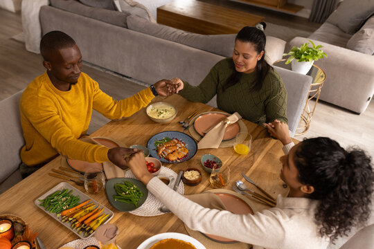 African American family holding hands around wooden dining table at home, sharing roasted chicken
