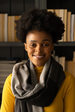 Woman smiling and wearing mustard ribbed turtleneck and knit gray scarf in home study with books