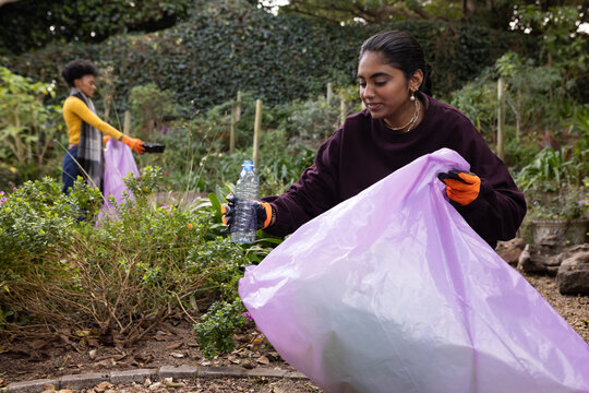 Diverse women picking litter in garden with orange gloves, placing bottle in purple bag, copy space