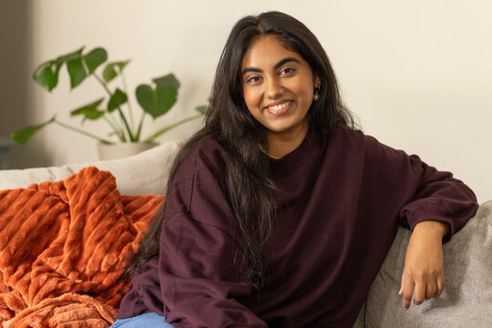 Indian woman smiling on sofa in living room wearing burgundy top, jeans, rust throw, plant, jewelry