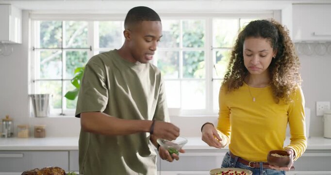 Couple finishing plating at kitchen island, woman sprinkling seeds on creamy dip, man placing roast
