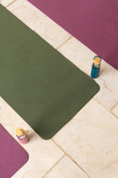 Olive-green yoga mat is occupying center on beige tiles, with magenta mats and teal pink bottles