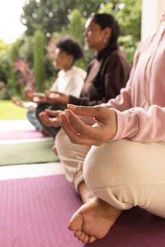 Diverse female friends meditating on yoga mats in garden, right woman wearing pink sweatshirt