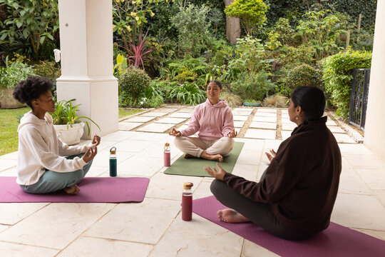 Diverse female friends meditating cross-legged on tiled patio with yoga mats and water bottles