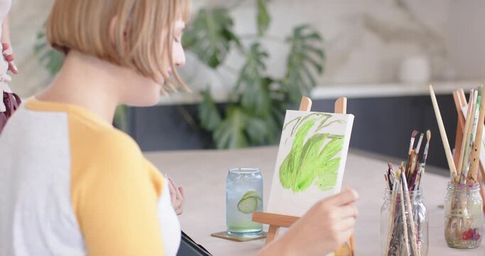 Female coach guiding seated woman painter painting leaves at easel in studio checking jar to refine