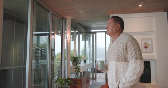 Senior Chinese man checking phone, picking mug, walking to balcony, sipping, watching misty trees