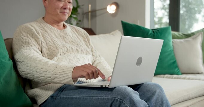Senior Chinese man seeing closed silver laptop, opening and typing, working on sofa wearing sweater