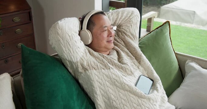 Senior Chinese man waking on couch by window, in sweater, headphones, reaching for phone, scrolling