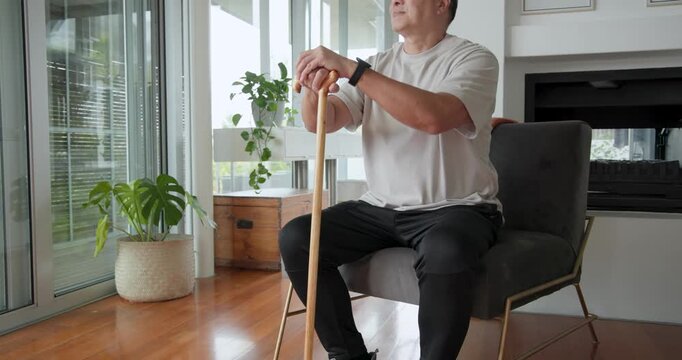 Senior Chinese man gripping cane in living room looking out doors pushing up walking toward balcony
