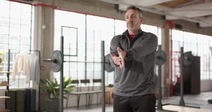 Senior male in loft gym warming for workout, moving from stretch to torso rotation near barbell