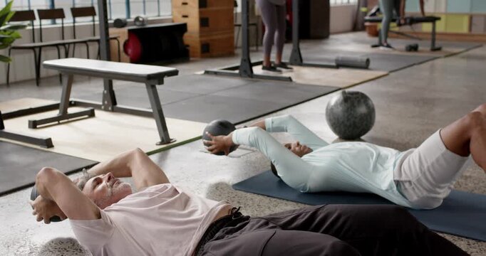 Diverse men lying supine with black med balls, performing sit-up-to-press reps in gym building core