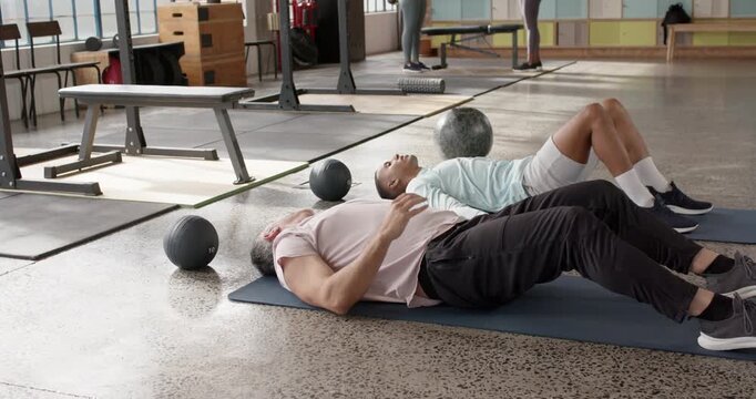 Diverse friends lying on blue mats in gym, man in pink polo raising smartphone, signaling partner