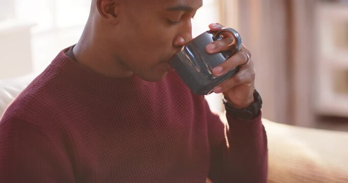 Man in maroon sweater lifting dark ceramic mug, inhaling aroma, savoring sip on couch by window
