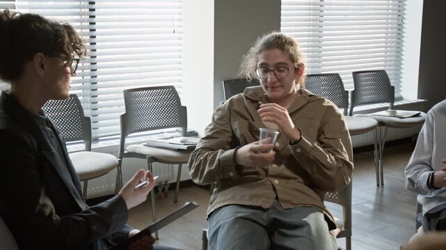Caucasian teenager with glasses drinking water and listening to female psychotherapist during group therapy session. Clip for mental health and counseling service campaigns