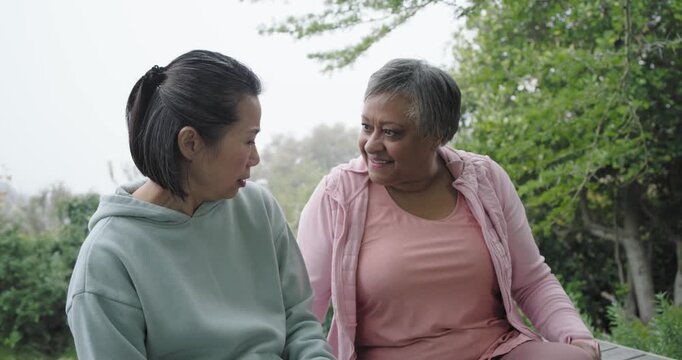 Diverse women sharing news touching knees laughing on wood bench in park wearing hoodies smartwatch