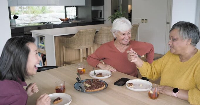 Diverse female friends sharing pecan pie bites, laughing, toasting glasses at home with smartphone