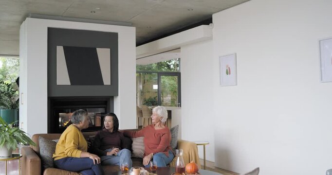 Mature women friends chatting as yellow-sweater woman gesturing on sofa at home, fireplace lighting