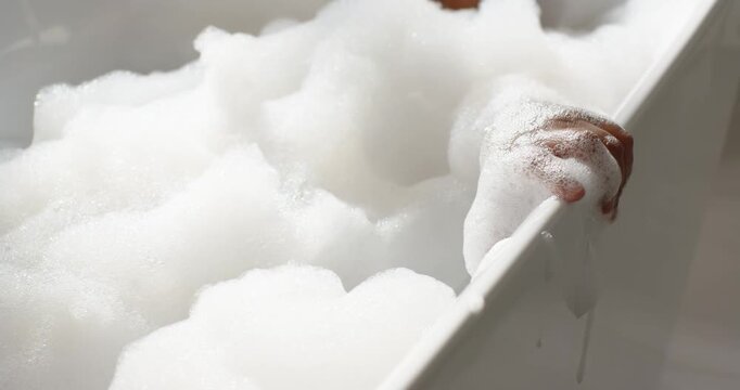 Hand is shifting along bathtub rim, causing foam to settle and reveal chin for relaxing