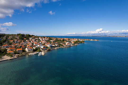 Drone shot capturing the beautiful Adriatic sea coast looking towards Poljana village from Preko side on Ugljan island, Croatia. Red-roofed houses line the turquoise water bay under a blue sky.