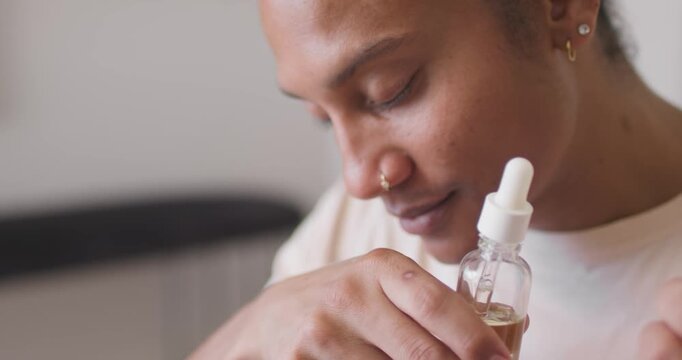 African American man holding dropper inspecting, dispensing skin oil to hand at home with nose ring