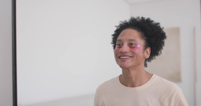 African American man standing by mirror applying pink under-eye patches for skincare, copy space