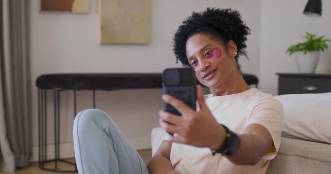 African American woman smiling, raising phone and angling for selfie on sofa wearing gel patches