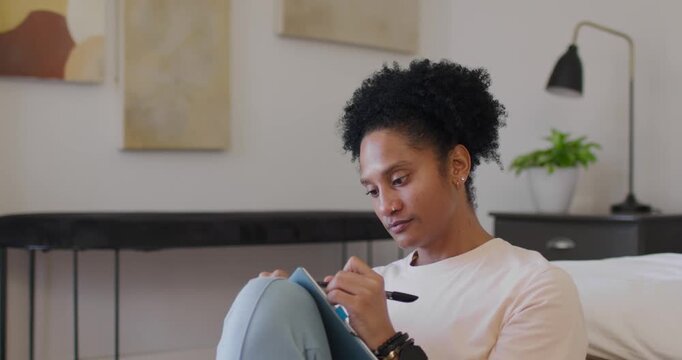 Adult African-American woman leaning in, holding pen, writing in blue notebook on sofa, reflecting