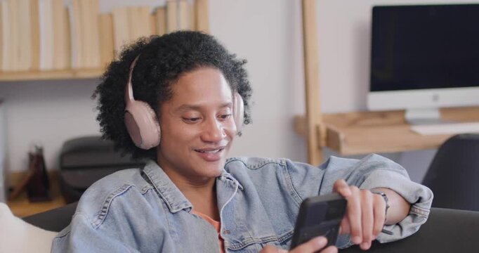 African man looking down at phone, scrolling, tapping, relaxing on couch at home with pink headset