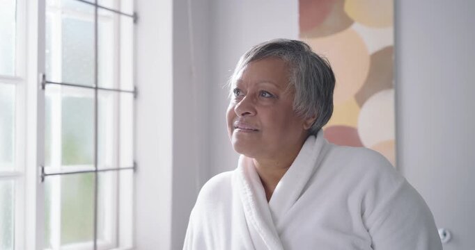 Sr African-American woman seeing light at window tilting, smiling in white robe near abstract art