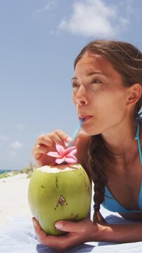 Happy beach bikini woman relaxing drinking fresh coconut water lying down sunbathing on fun Caribbean vacation tanning under the tropical sun. Asian model enjoying suntan. Vertical video/