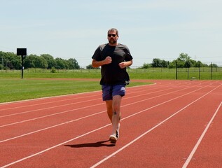 Fototapeta premium Adult man preparing for outdoor workout adjusting wireless earbuds stretching legs on railing and running on a red running track at a local stadium