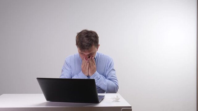 Man in a blue shirt blowing his nose with a tissue while working on a laptop at a white desk, illustrating illness, allergy, and sick leave in a work environment