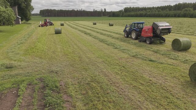 Blue tractor pulls a round baler producing a fresh hay bale in a rural field, with cut grass, forest background, and summer farming atmosphere.