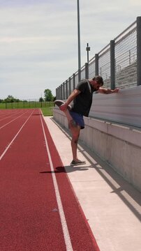 Adult man preparing for outdoor workout adjusting wireless earbuds stretching legs on railing and running on a red running track at a local stadium