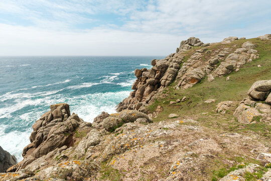 View of the rugged granite cliffs and turquoise waters at Nanjizal Bay with grassy slopes under a bright sky in Saint Just, England, United Kingdom.