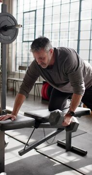 Vertical video: Midlife man placing hand on bench in gym, lifting hex dumbbell, rowing for strength