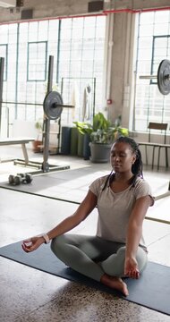 Vertical video: African American woman sitting on mat in gym, closing eyes, breathing to focus
