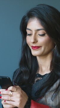 Elegant close up portrait Asian woman holding smartphone and smiling softly. Female reading message and typing reply on mobile. Indoor calm lifestyle moment. Young Indian lady texting on phone.